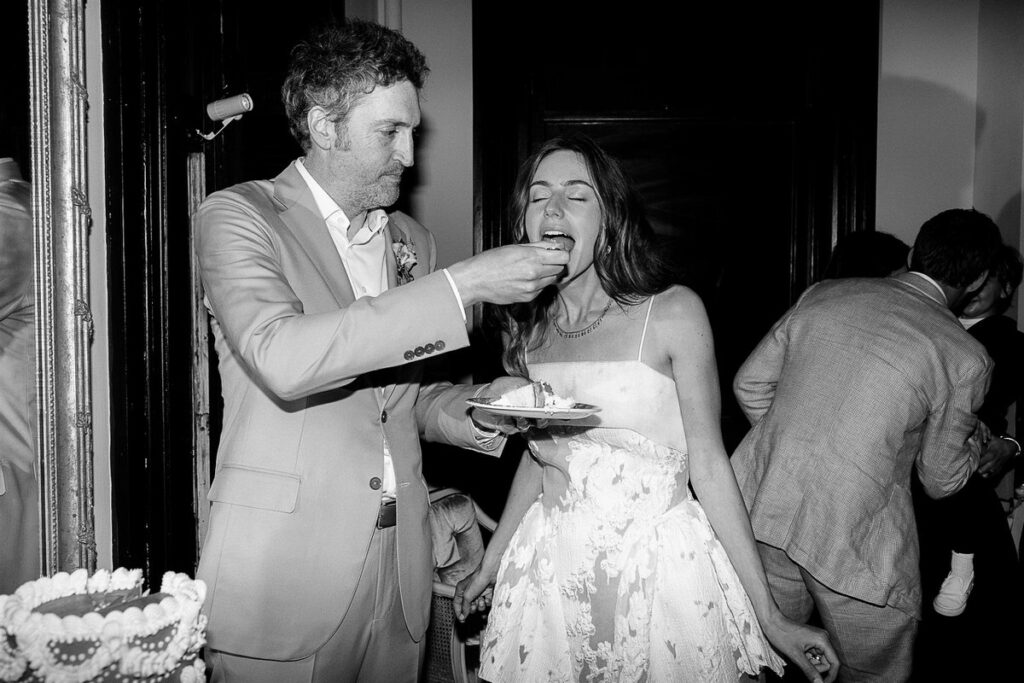 The groom feeds the bride a piece of wedding cake, both laughing.
