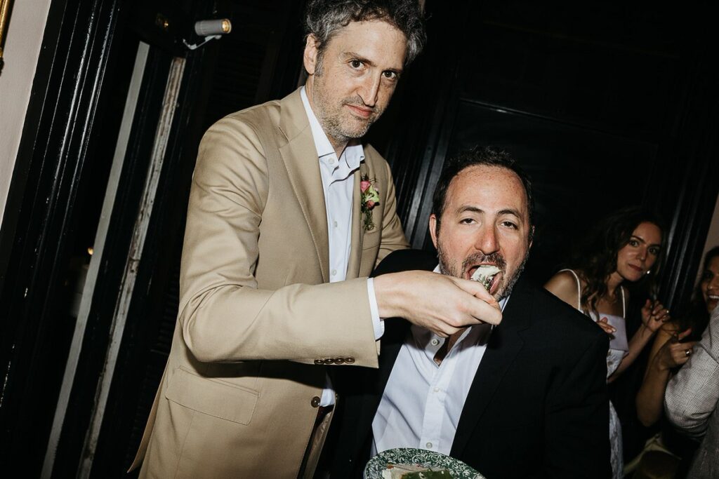 The groom feeds a piece of cake to a male guest, captured in a candid moment.
