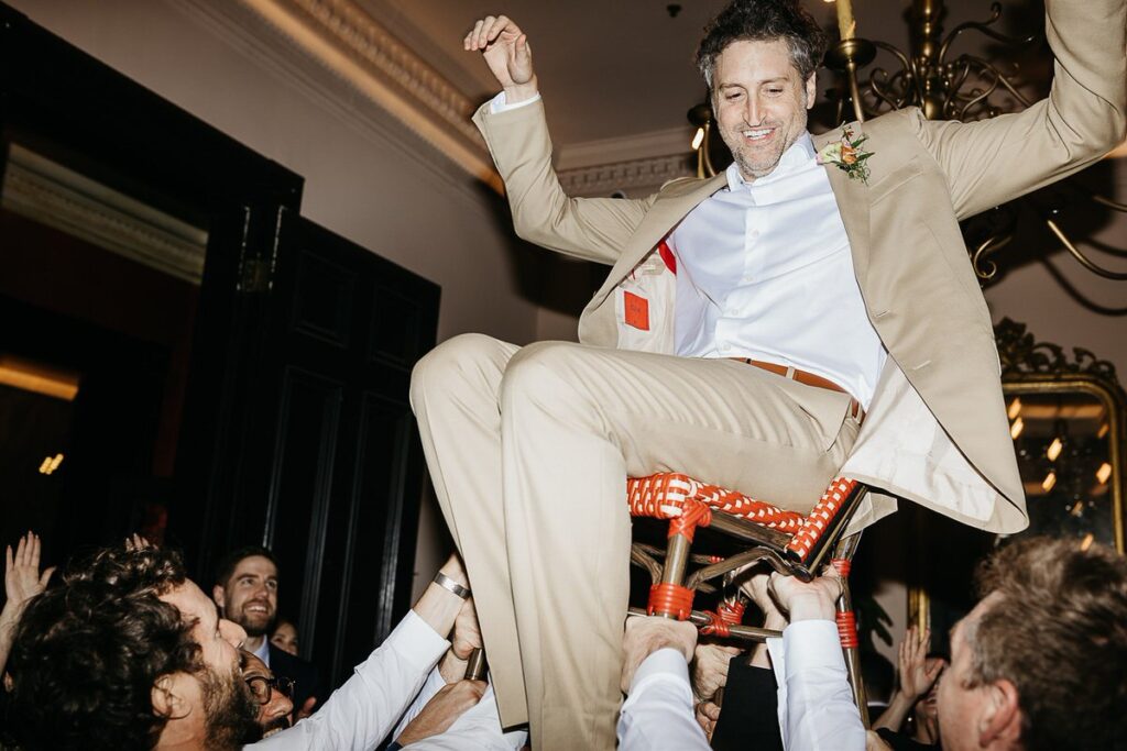 The groom is lifted in a chair during the traditional Jewish hora dance, smiling broadly.
