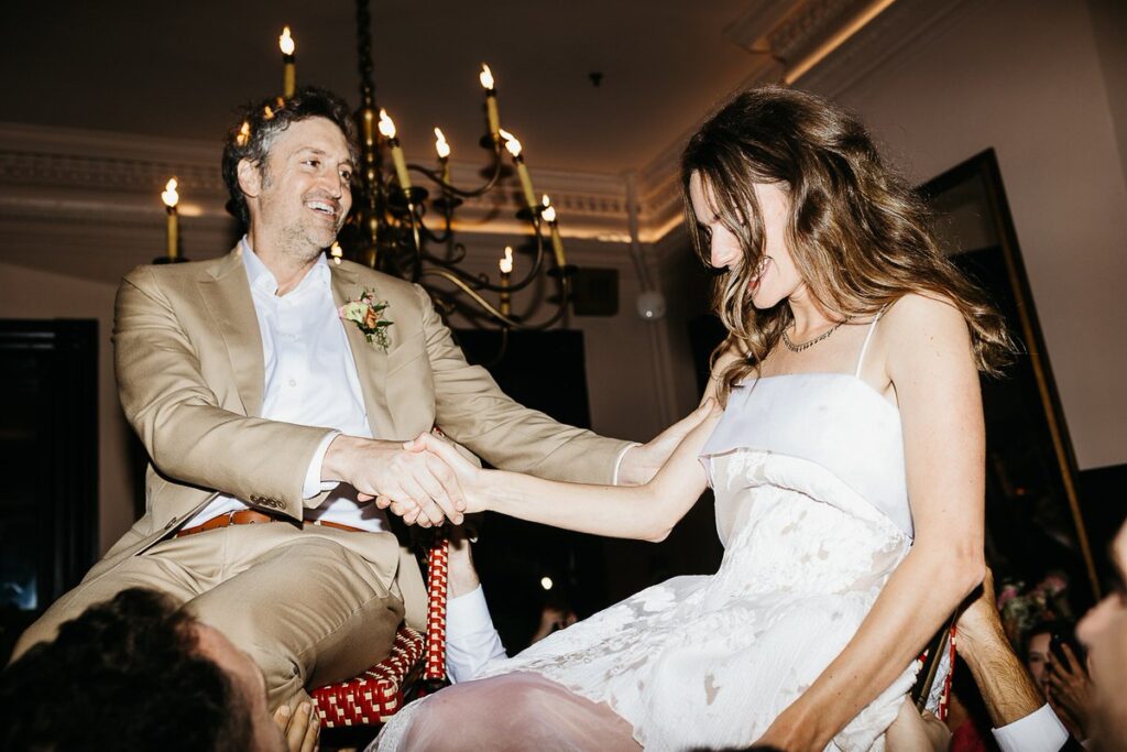 The bride and groom is lifted into the air on a chair during the Hora dance, smiling and holding the bride's hand.

