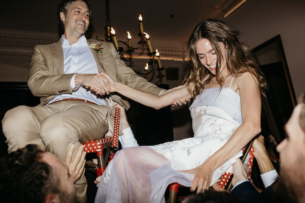 The bride and groom hold hands and smile as they are lifted into the air on chairs during the Hora dance.

