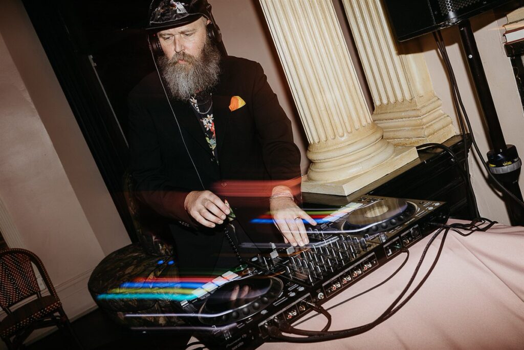 A DJ with a full beard and black hat mixes music on a turntable, set against a backdrop of classic columns.

