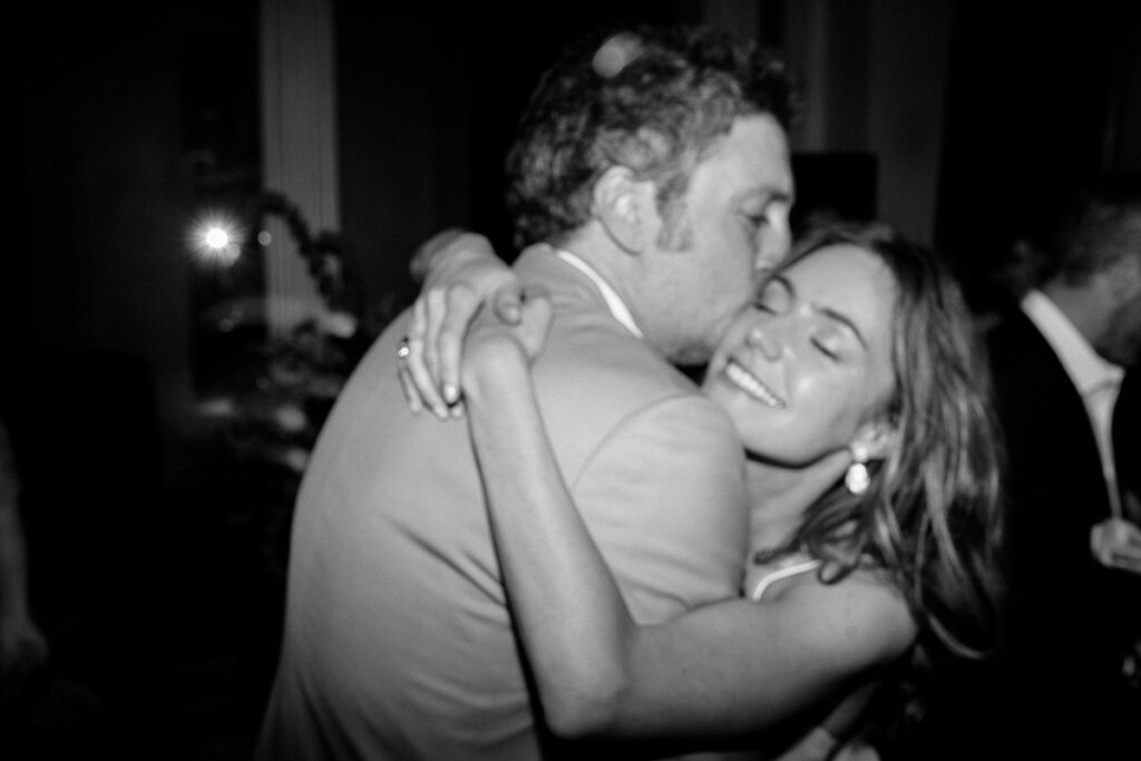 A black-and-white photo of the groom kissing the bride's cheek as they embrace during the reception.

