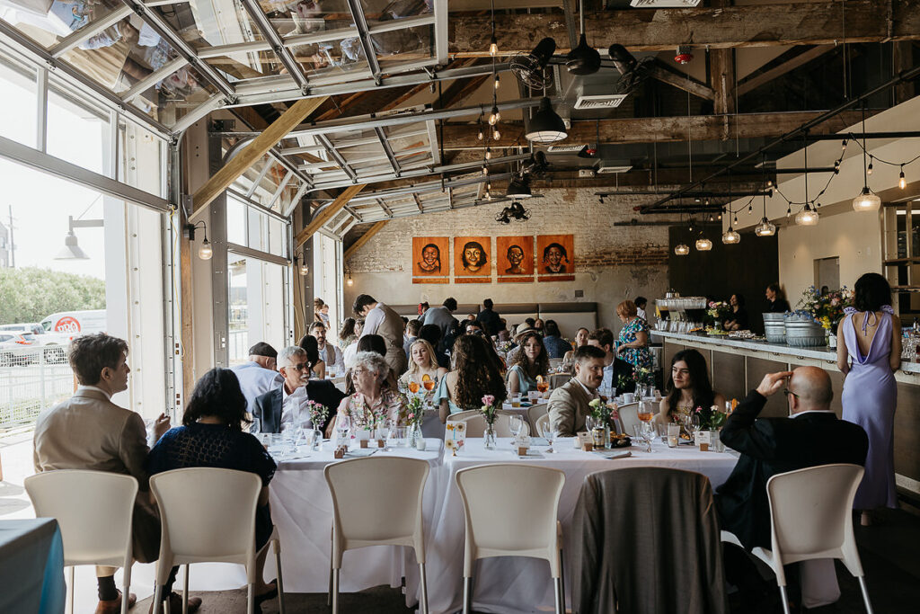 A brunch setting inside a rustic venue with guests seated at a long table, engaged in conversation. 