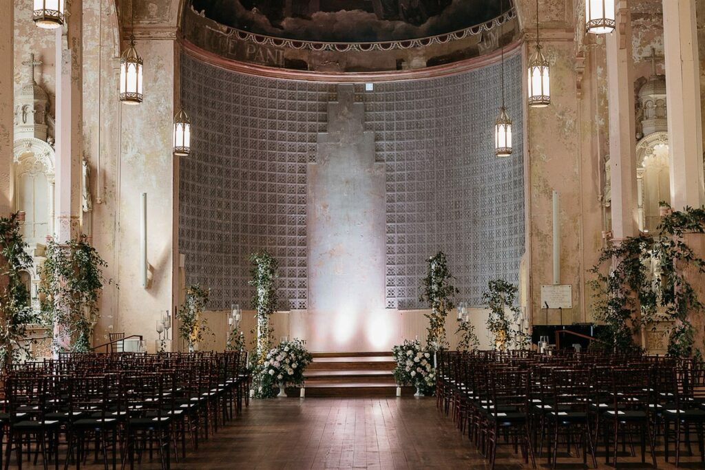 A wide view of a historic venue decorated for a wedding ceremony, with rows of chairs and a beautifully adorned altar.
