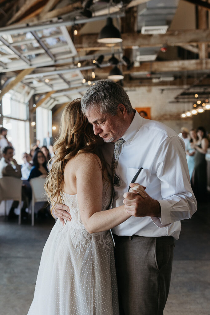 Bride dancing with her father at the reception, both smiling and close, with guests and rustic decor in the background