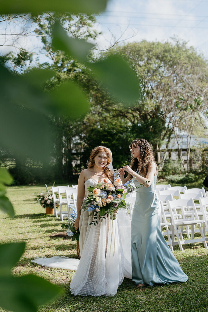 Bride smiling while bridesmaid adjusts her hair, surrounded by white chairs outdoors.
