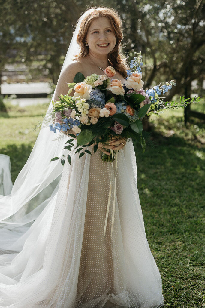 The bride standing outdoors, smiling and holding a vibrant bouquet of flowers