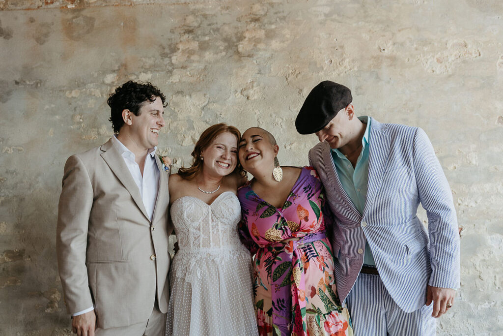 Bride and groom smiling with two friends, posing against a rustic wall.
