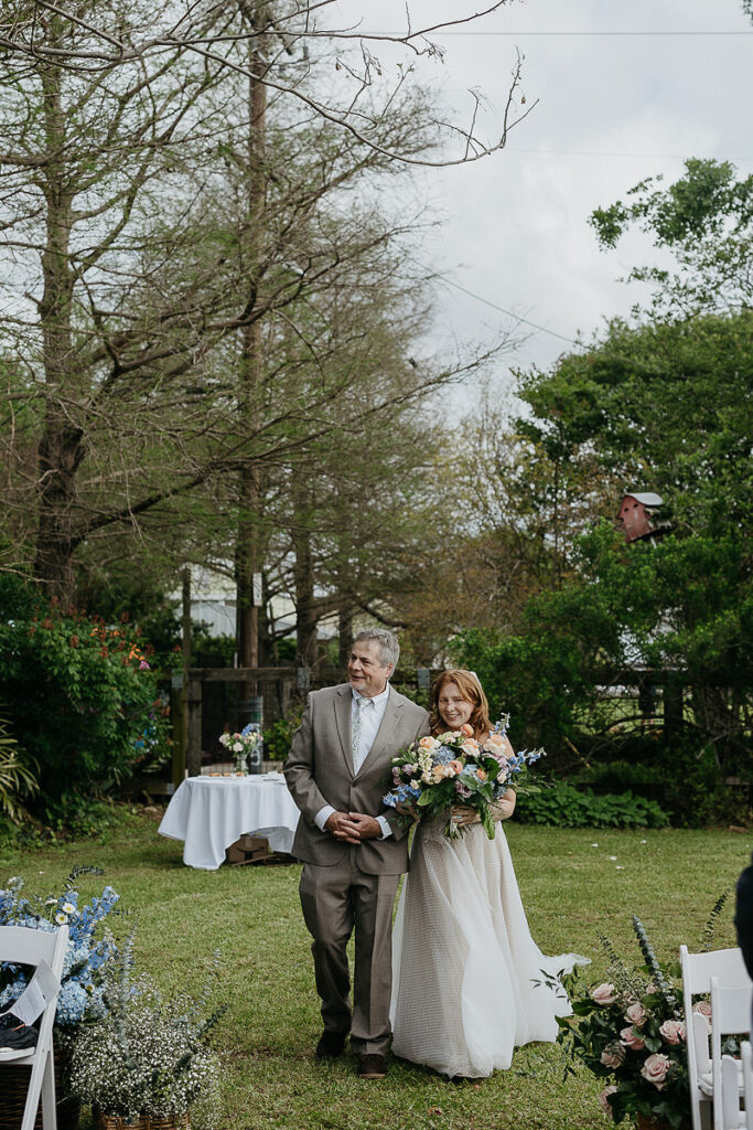 Bride, holding a large bouquet, walks down the aisle with her father in a lush outdoor setting