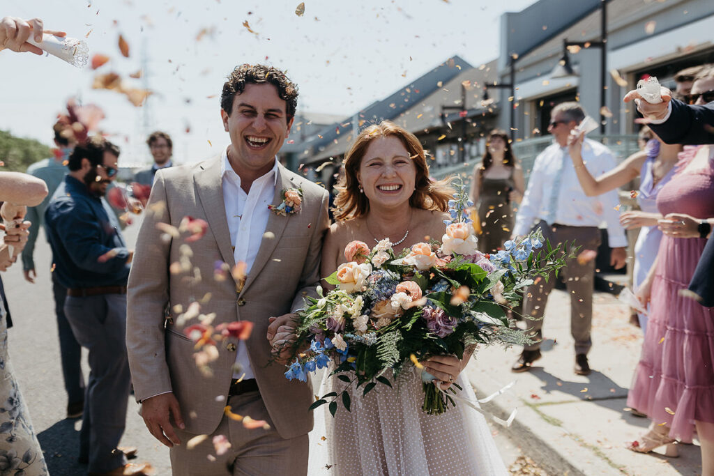 Bride and groom walk down the street, surrounded by guests throwing flower petals in celebration