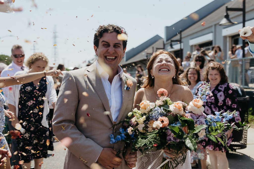 Bride and groom share a joyful moment with guests, smiling and laughing amidst flying flower petals