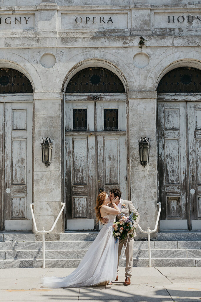 Bride and groom share a kiss in front of a historic opera house with rustic wooden doors