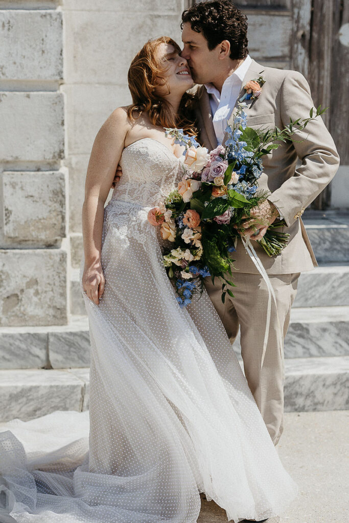 Bride and groom share a tender moment, smiling and embracing outside an old building with a bouquet of flowers