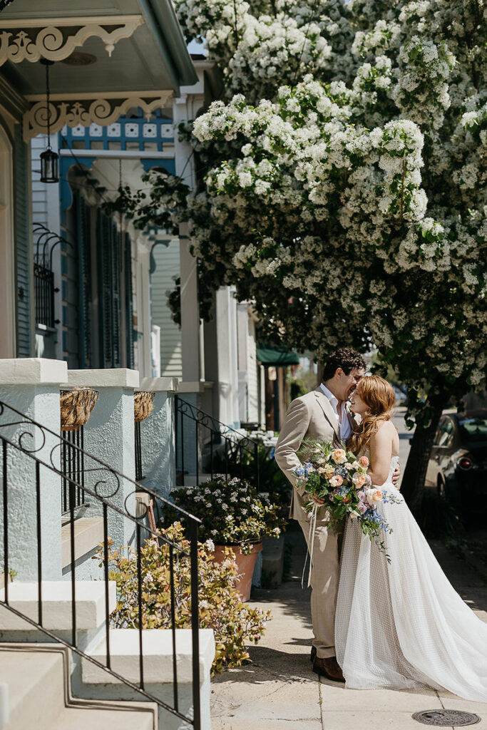 Bride and groom stand close together on a picturesque street, surrounded by blooming flowers and charming houses