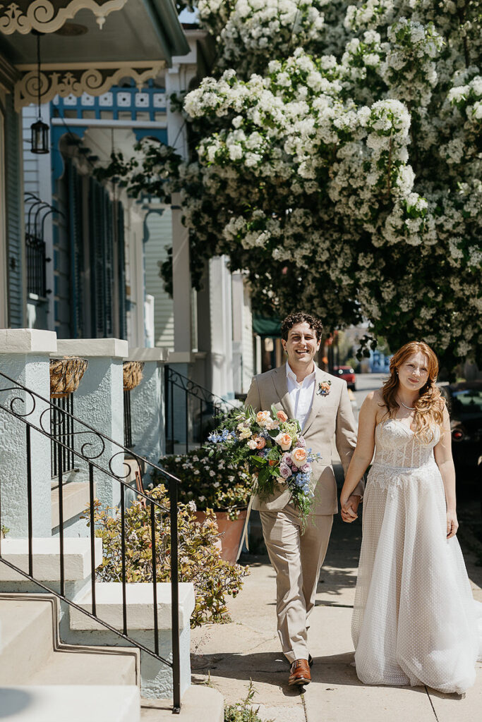 The bride and groom walk hand in hand on a sunny street, with the groom holding a vibrant bouquet of flowers