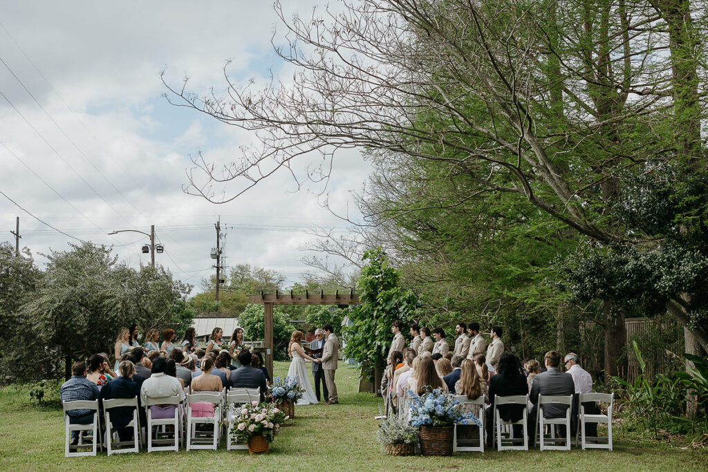 An outdoor wedding ceremony with guests seated on white chairs, as the bride and groom stand under a wooden pergola.
