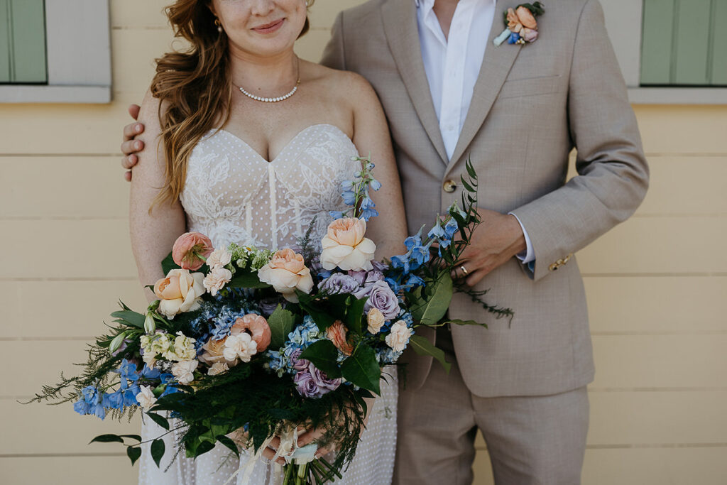 Close-up of the bride holding a large bouquet of pastel flowers, standing next to the groom in a beige suit.
