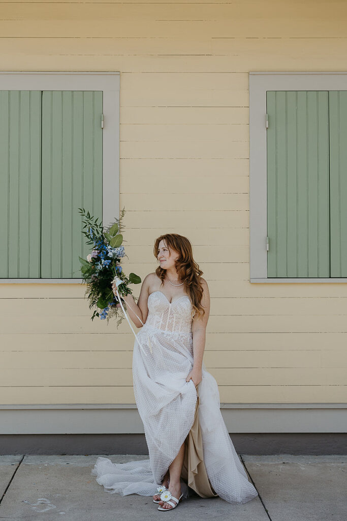 The bride poses alone in front of a yellow building with green shutters, holding her bouquet and smiling.
