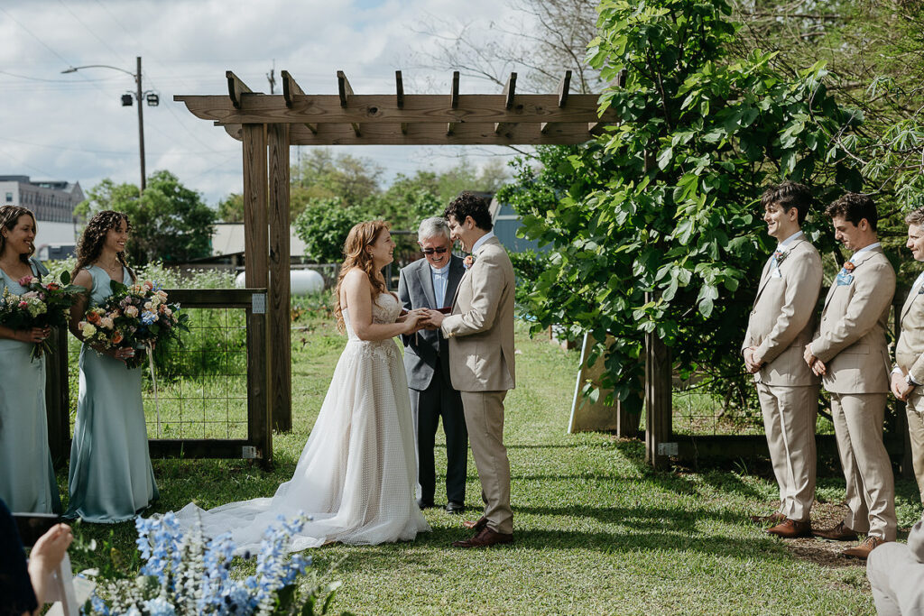 The bride and groom exchange rings under a wooden pergola during their outdoor wedding ceremony.
