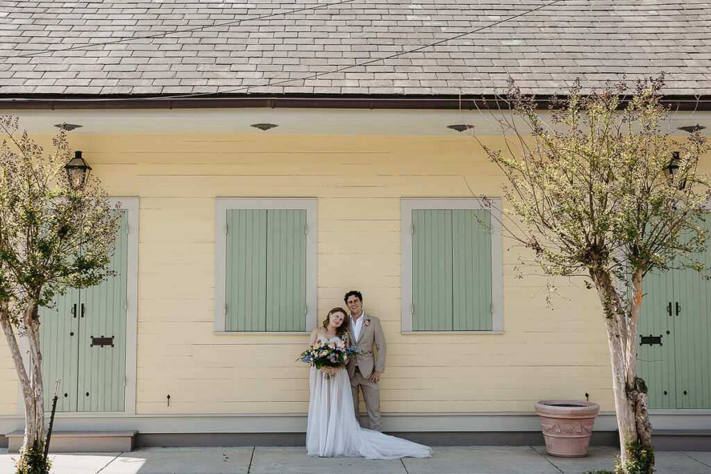 The bride and groom stand close together against a yellow building with green shutters, holding a large bouquet.
