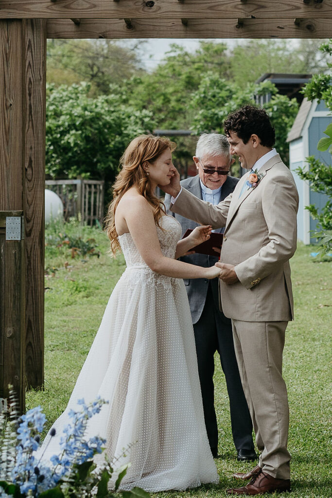 The groom gently touches the bride's face during their wedding ceremony under a wooden pergola.
