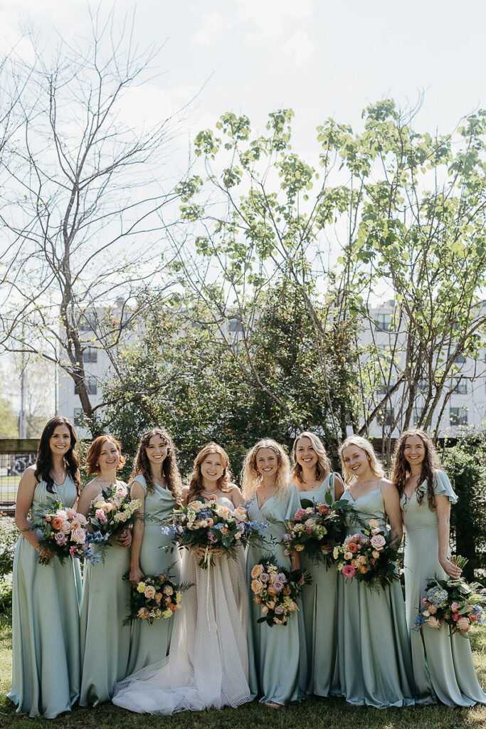 The bride stands with her bridesmaids, all dressed in light blue dresses, each holding a colorful bouquet.
