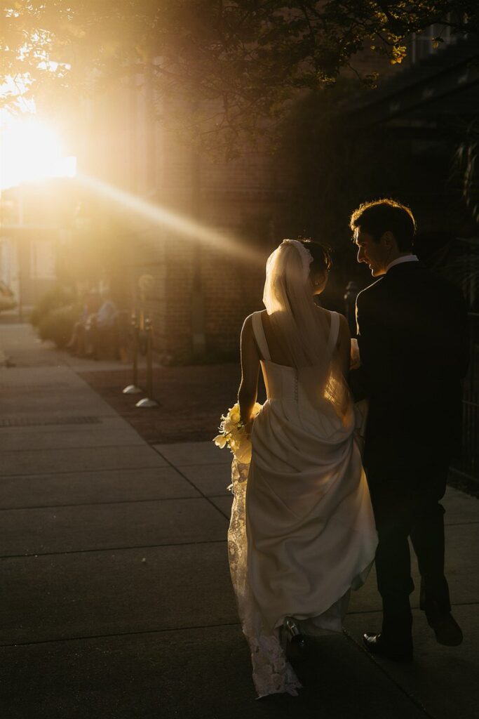 A bride and groom walking along a sidewalk at sunset 