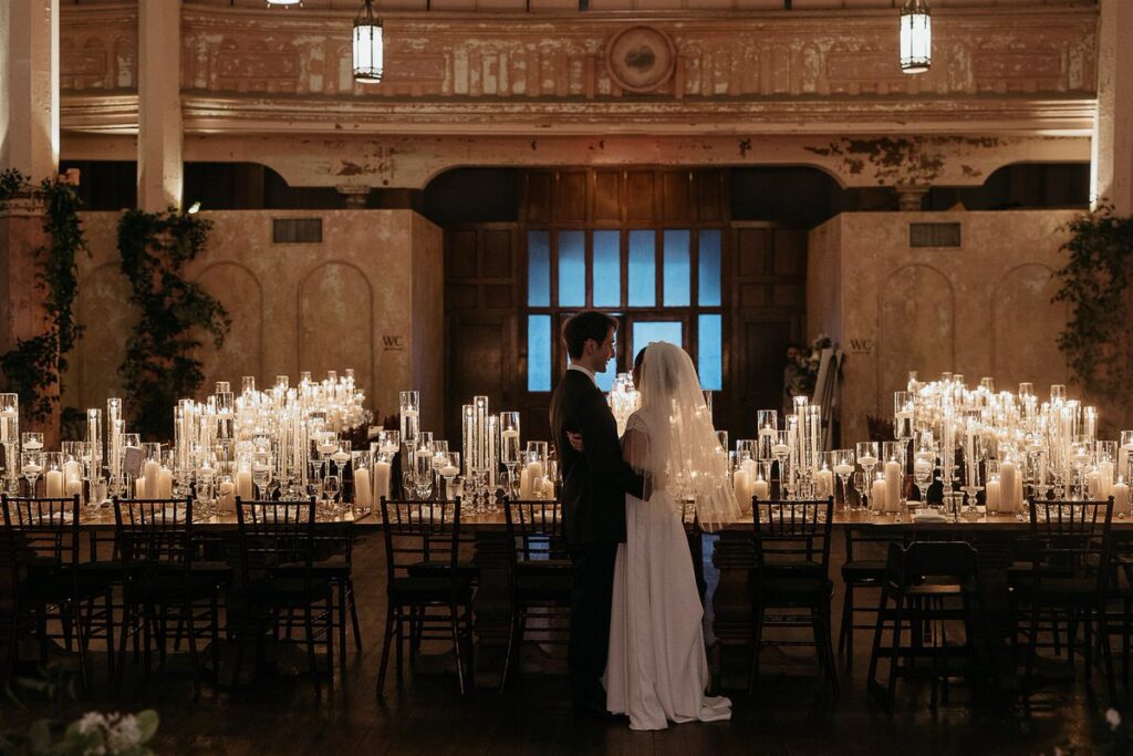 A bride and groom embracing while standing in front of reception tables 