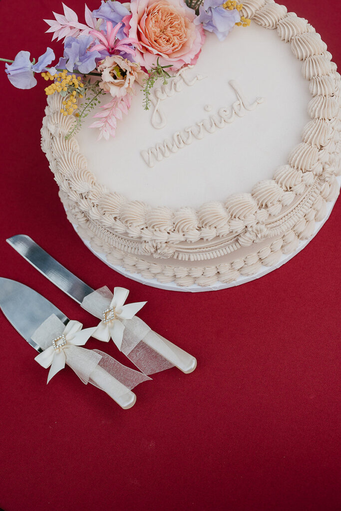 A wedding cake with floral decorations and the words "just married" on top, with a cake knife and server decorated with bows and flowers on the side.
