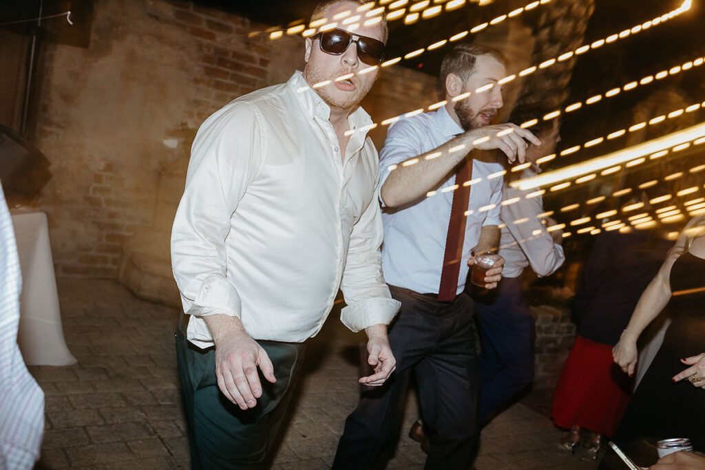 A man dancing at a wedding reception, with motion blur from string lights creating streaks of light in the background.
