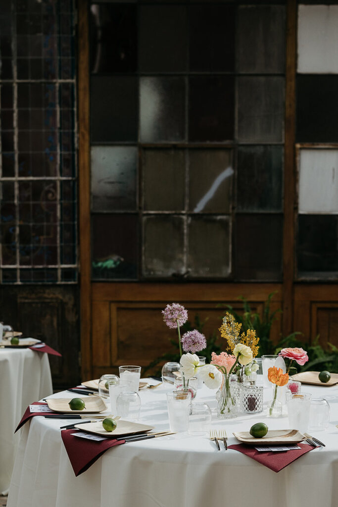 A wedding reception table with floral centerpieces and place settings, set against a backdrop of a vintage window with multiple panes.
