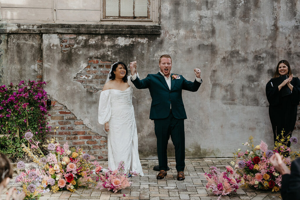 The bride and groom standing in front of an old brick wall, cheering with their hands raised, surrounded by colorful floral arrangements.
