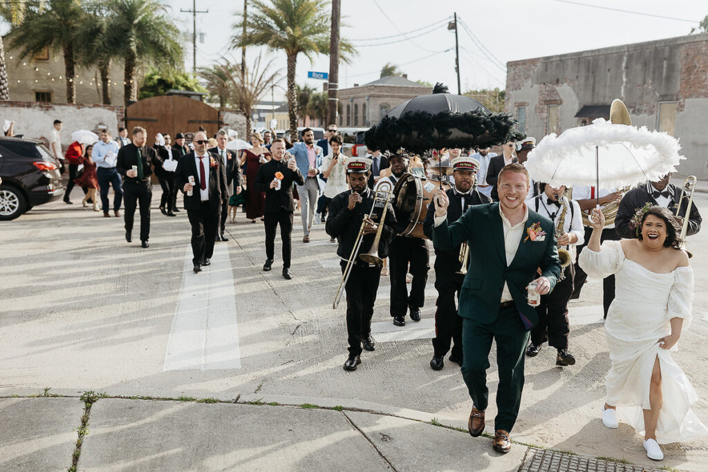 A wedding parade with a brass band and guests walking down the street, led by the bride and groom holding feathered umbrellas.
