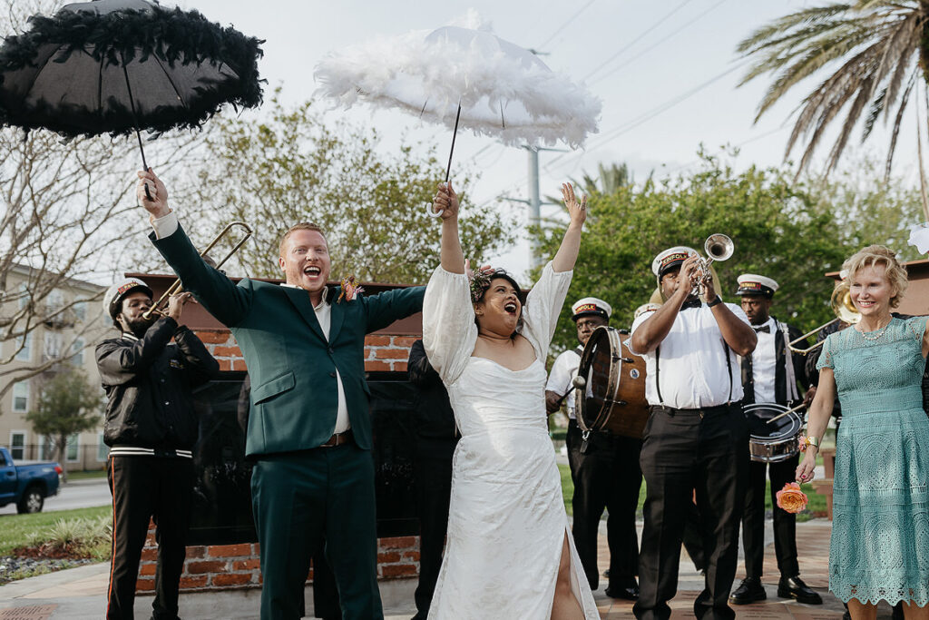 The bride and groom holding feathered umbrellas and cheering in front of a brass band.
