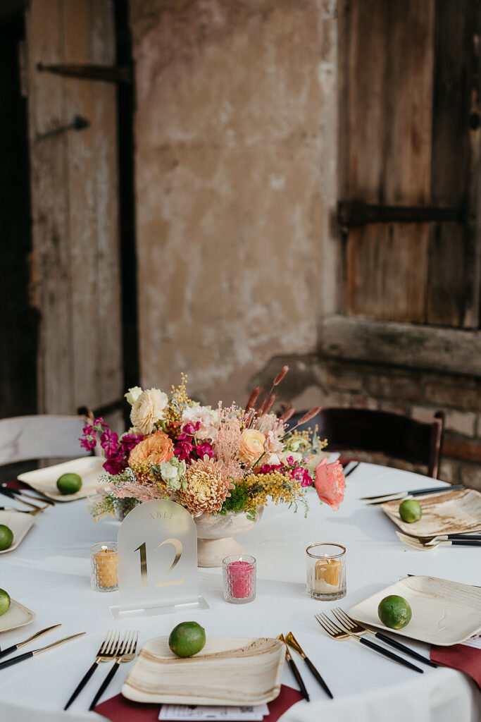 Close-up of a wedding reception table with a floral centerpiece, candles, and place settings including limes on wooden plates.
