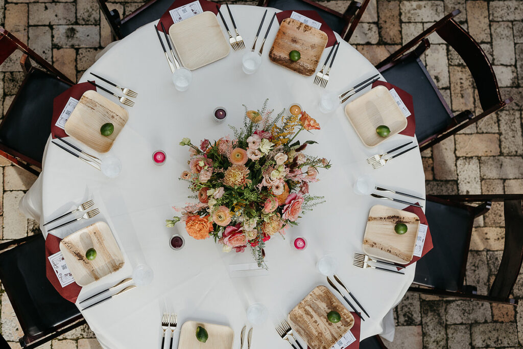 A round table set for a wedding reception, viewed from above, with floral centerpieces and place settings that include wooden plates and limes.
