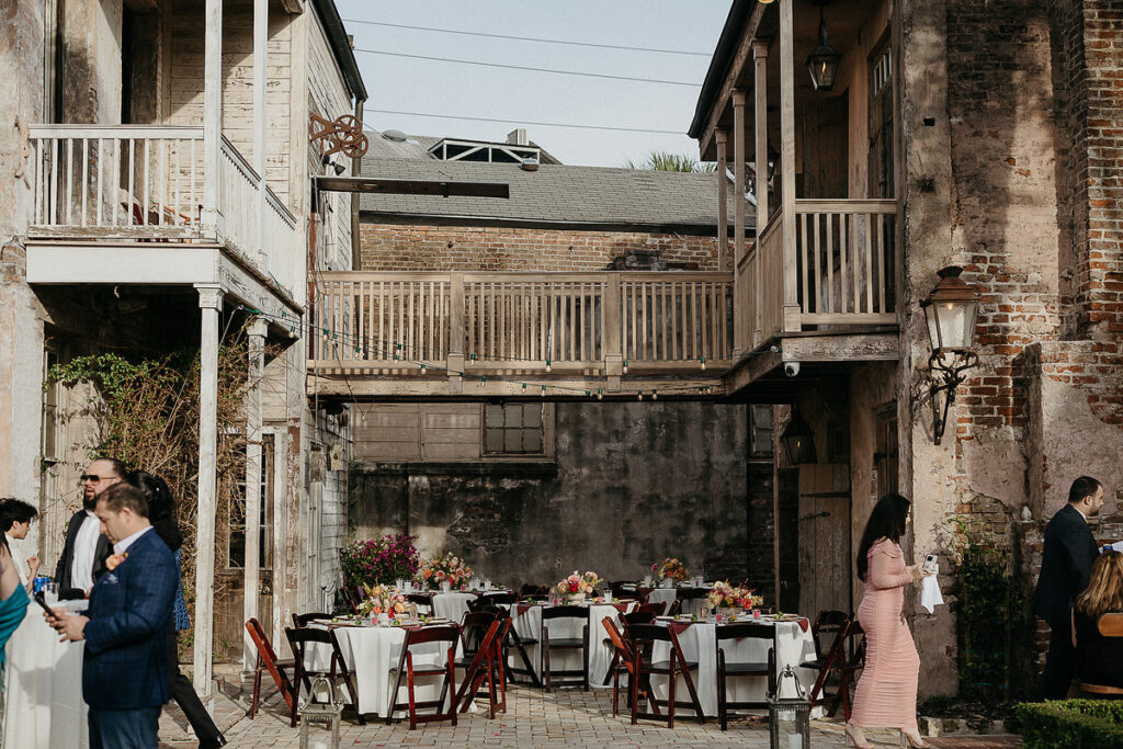 An outdoor courtyard setting with round tables covered in white tablecloths and floral centerpieces, surrounded by rustic buildings.
