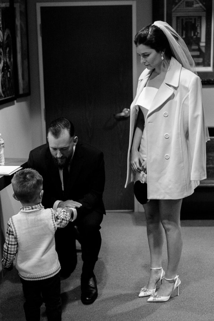 A groom kneeling down holding the hand of a small child with a bride standing next to them 
