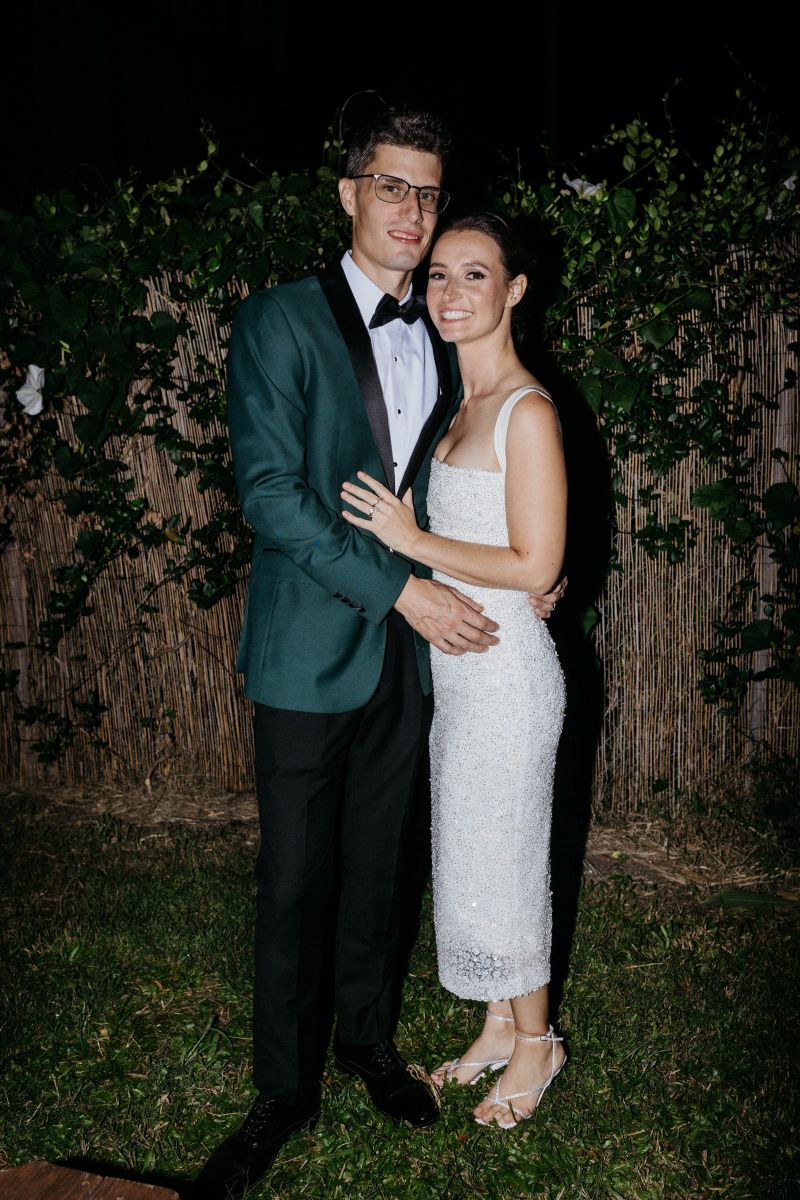 a couple hugging each other and smiling the woman is wearing a white dress the man is wearing a green dress jacket and black pants 