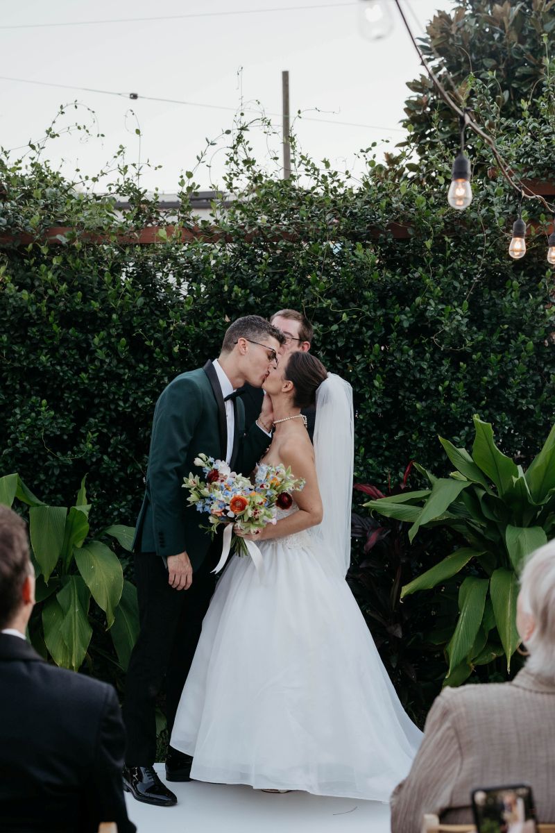 a copule kissing at their wedding ceremony the woman is wearing a white veil and dress and holding a bouquet of flowers the man is wearing a green suit behind them is their officiant and a garden