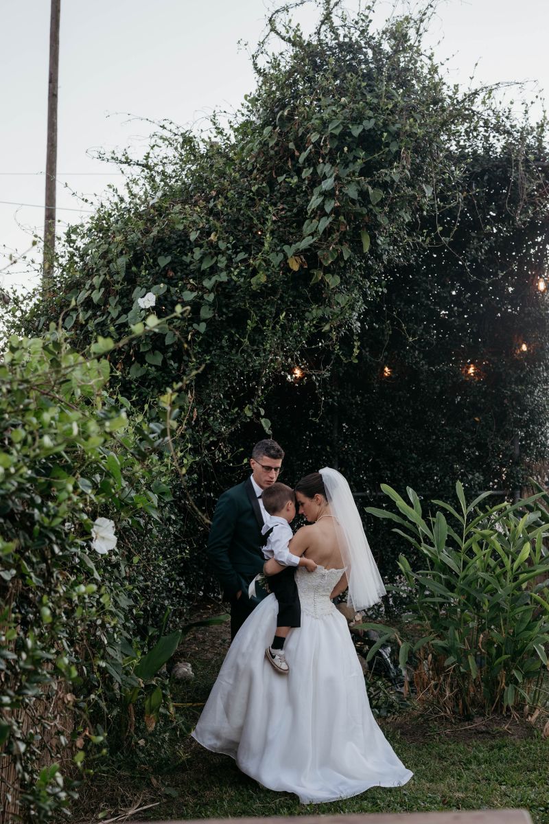 a couple in a graden the woman is wearing a white wedding dress and veil and is holding her young son her partner is standing in front of them 