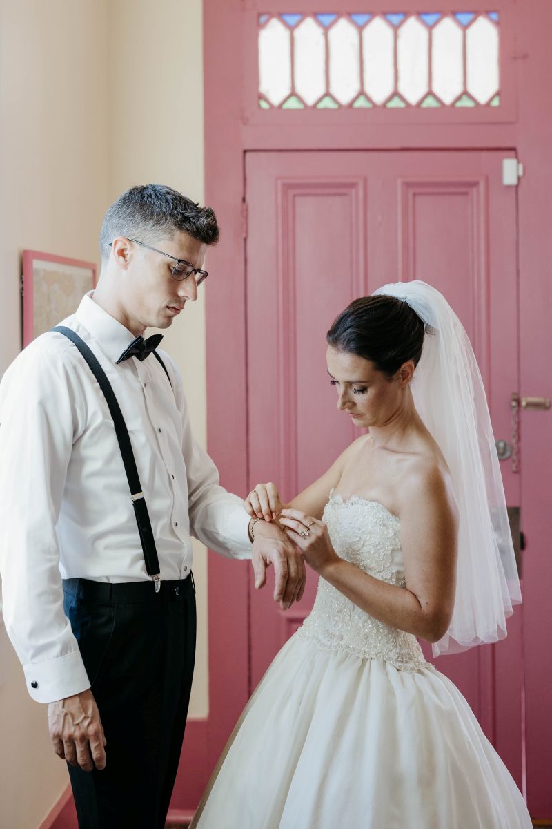 a woman is a white wedding dress and veil is fixing her partner's cuff on his shirt they are both standing next to a pink door 