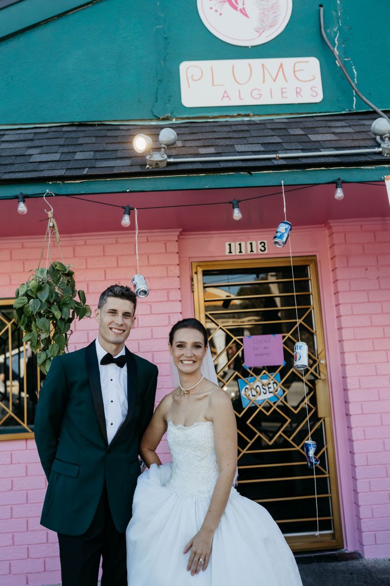 a couple standing in front of their restaurant the woman is wearing a white wedding dress and veil the man is wearing a suit and they are both smiling 