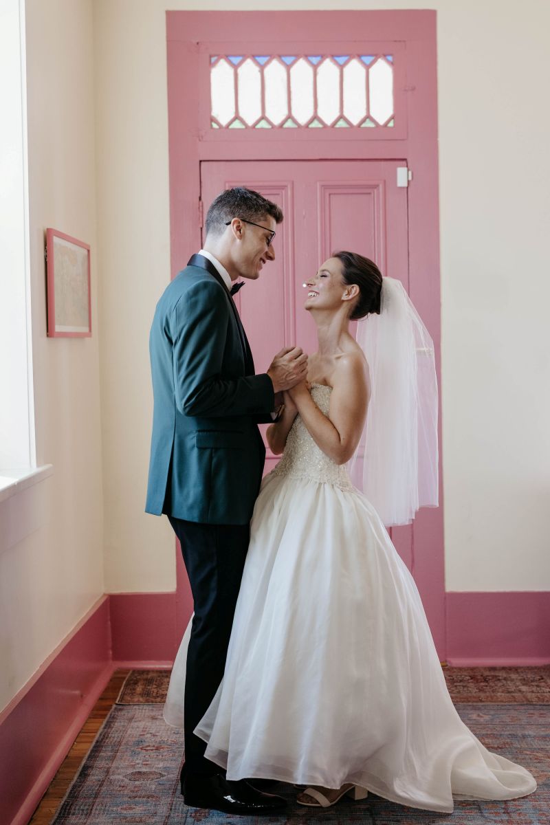 a woman and a man are both standing in a room next to a pink door and smiling at each other the woman is wearing a white wedding dress and veil and the man is wearing a blue suit 