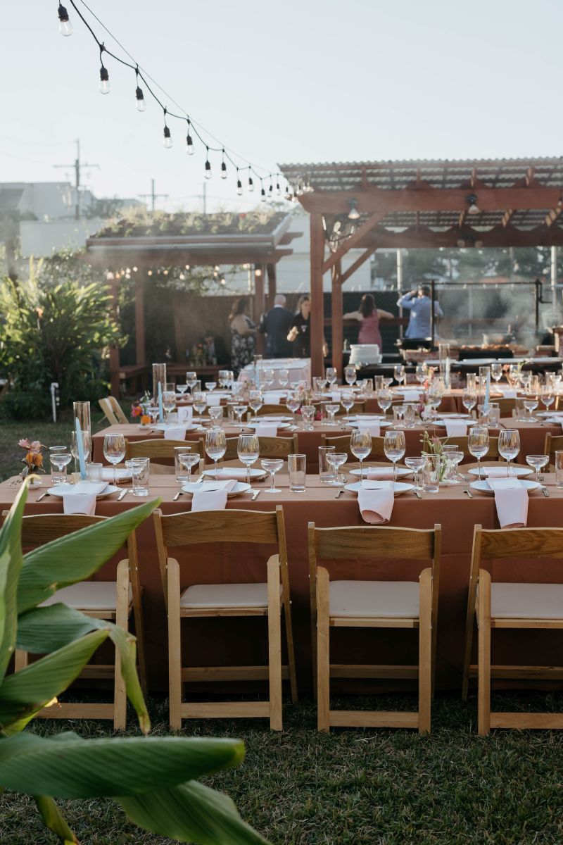 tables with tableclothes, chairs, plates, vases, and napkins on display there is also an outdoor grill and string lights overhead 