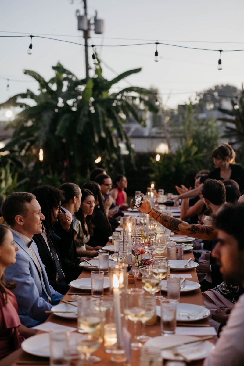 a long banquet table with wedding guests above them are string lights on the table are candles, plates, and glasses 