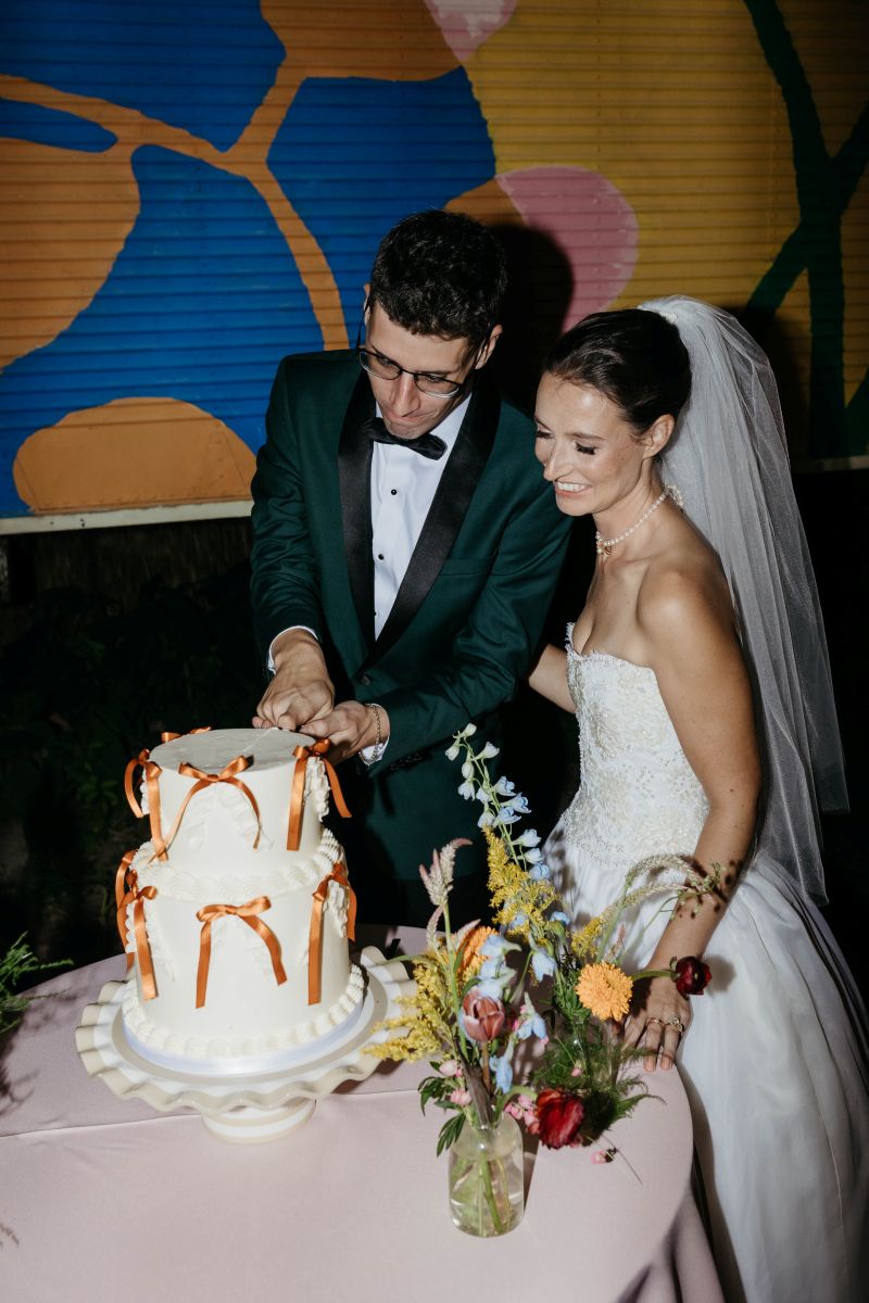 a couple cutting their cake at their wedding the man is cutting the cake and the woman is watching and smiling she is wearing a white wedding dress and veil 