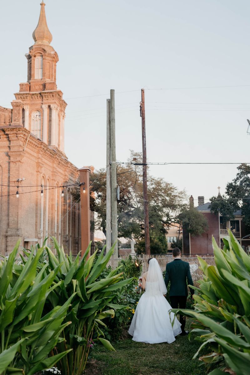 a couple walking hand in hand throuhg a garden in front of them is a church the woman is wearing a white wedding dress and veil the man is wearing a suit 