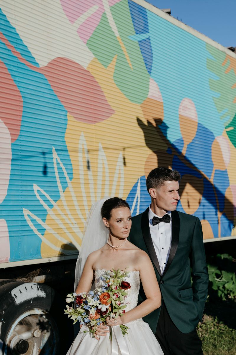 a couple standing next to each other behind them is a trailer truck with an array of colors and designs the woman is wearing a white wedding dress and veil and holding a bouquet of flower sthe man is wearing a suit they are boht looking off to the side 